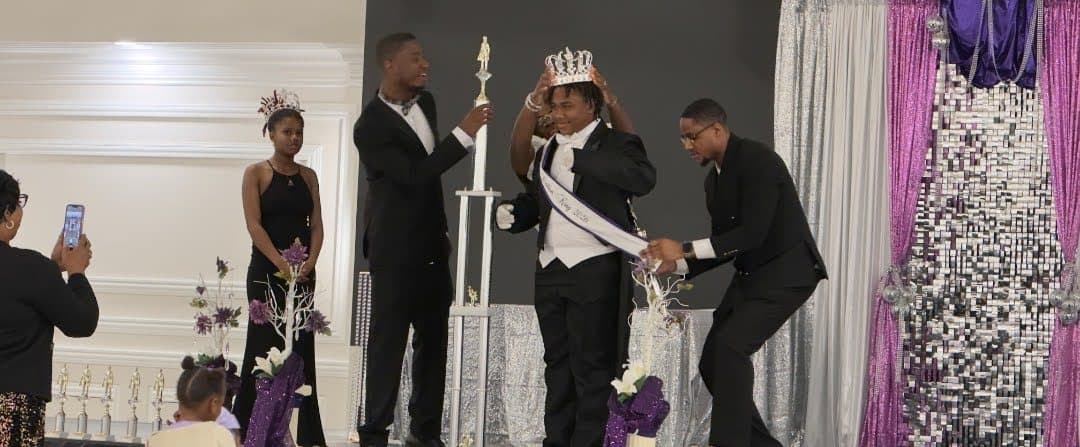 A young gentleman is crowned on stage beside a trophy