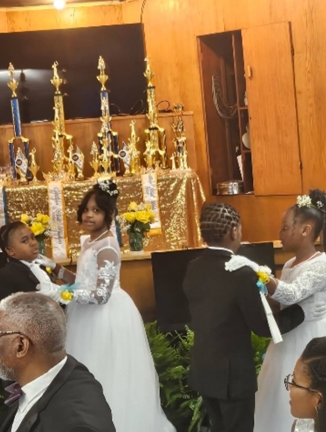 Young ladies in white gowns and young gentlemen in tuxedos process past a table of trophies