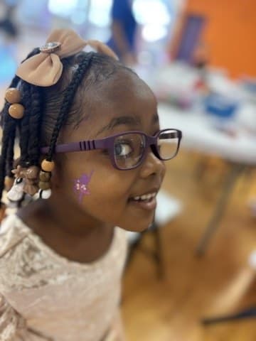 A girl in glasses smiling over her shoulder at the iKIDZ craft table